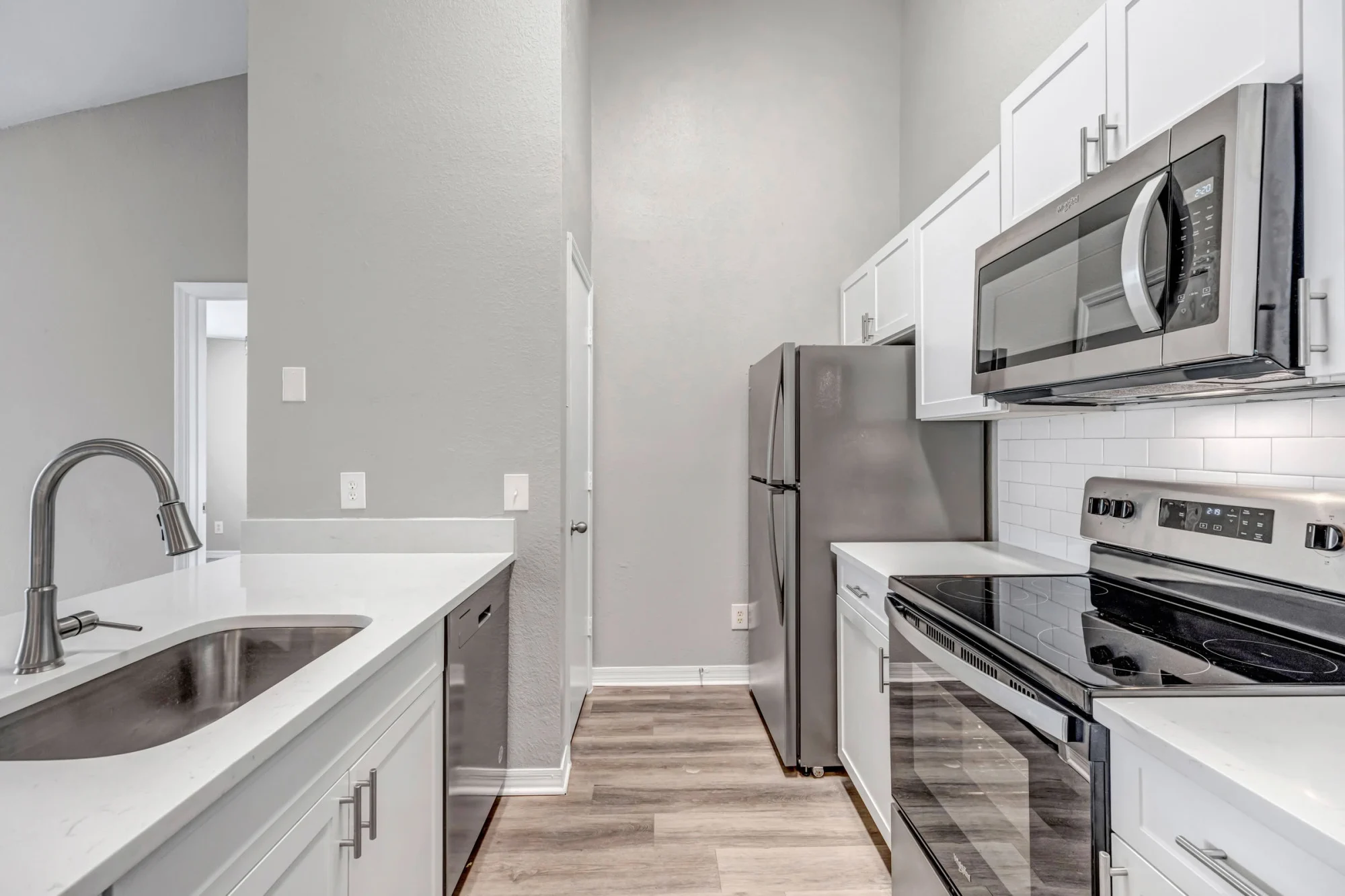Galley-style apartment kitchen with white cabinets, quartz countertops, stainless steel appliances, white subway tile backsplash, deep stainless sink, and light wood-look flooring.